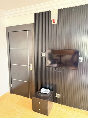 A room with a closed door, a flat-screen TV mounted on a dark wall, and a small wooden chest of drawers. - Floyd Msquare Hotel (Lagos)