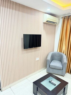 A modern living room with a flat-screen TV mounted on a textured wall, a grey armchair, and a glass-top coffee table with a remote control. - Floyd Msquare Hotel (Lagos)