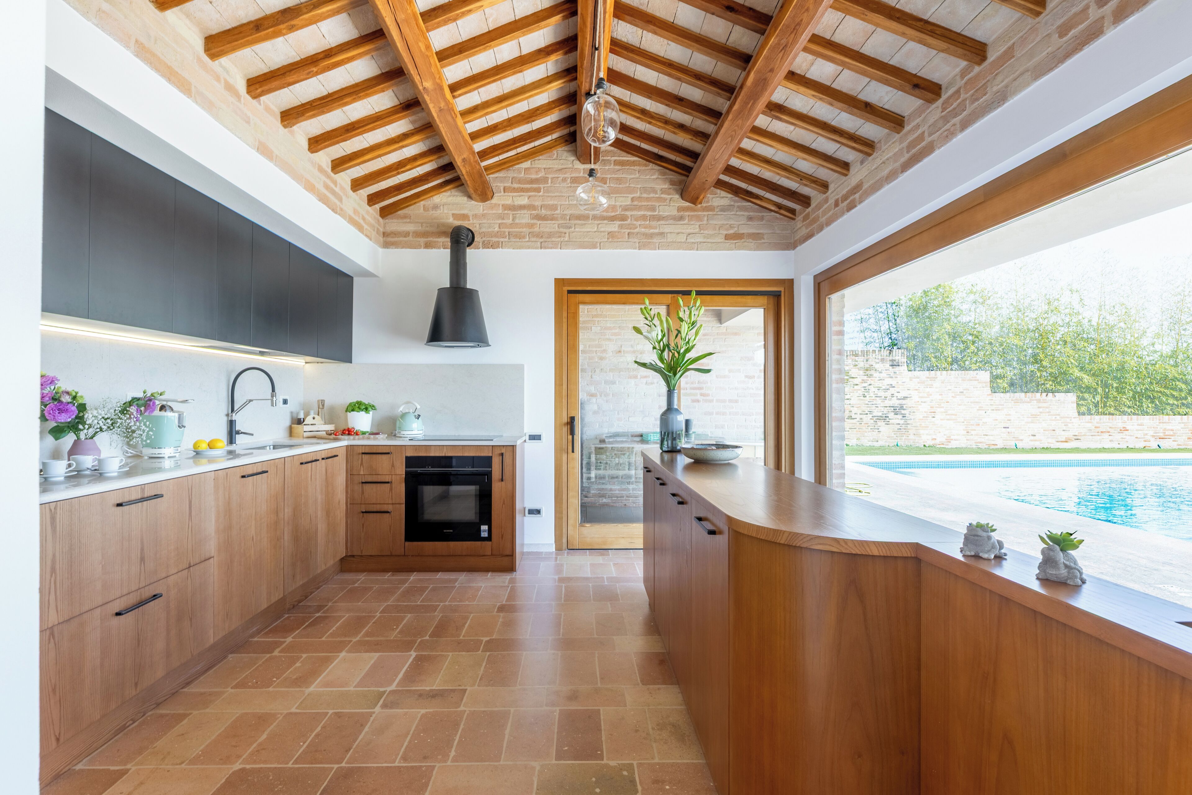 A modern kitchen with wooden cabinets, a built-in oven, and a large window overlooking a swimming pool.