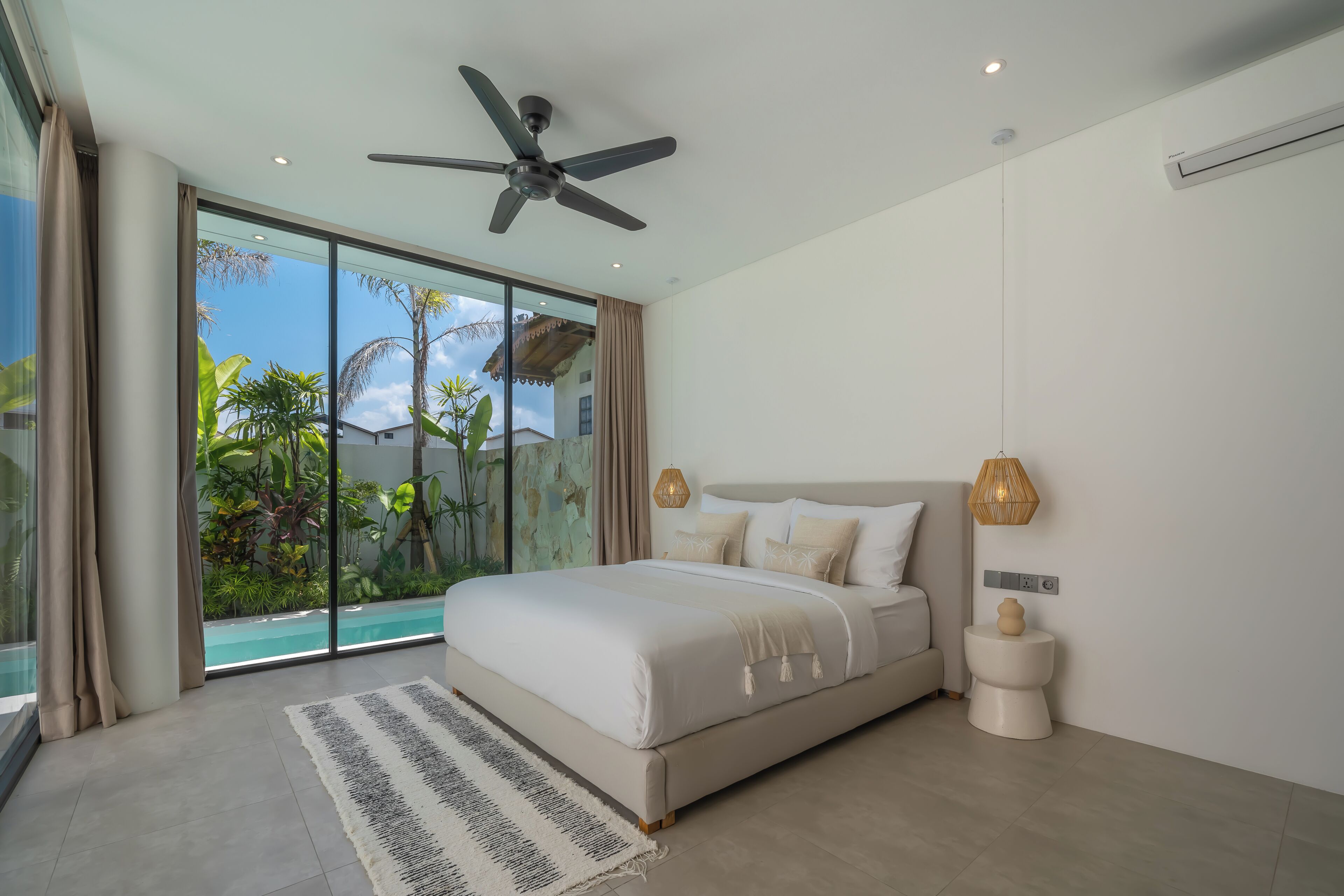 Une chambre avec un lit, un ventilateur de plafond, une grande fenêtre donnant sur un paysage verdoyant et un climatiseur fixé au mur.
