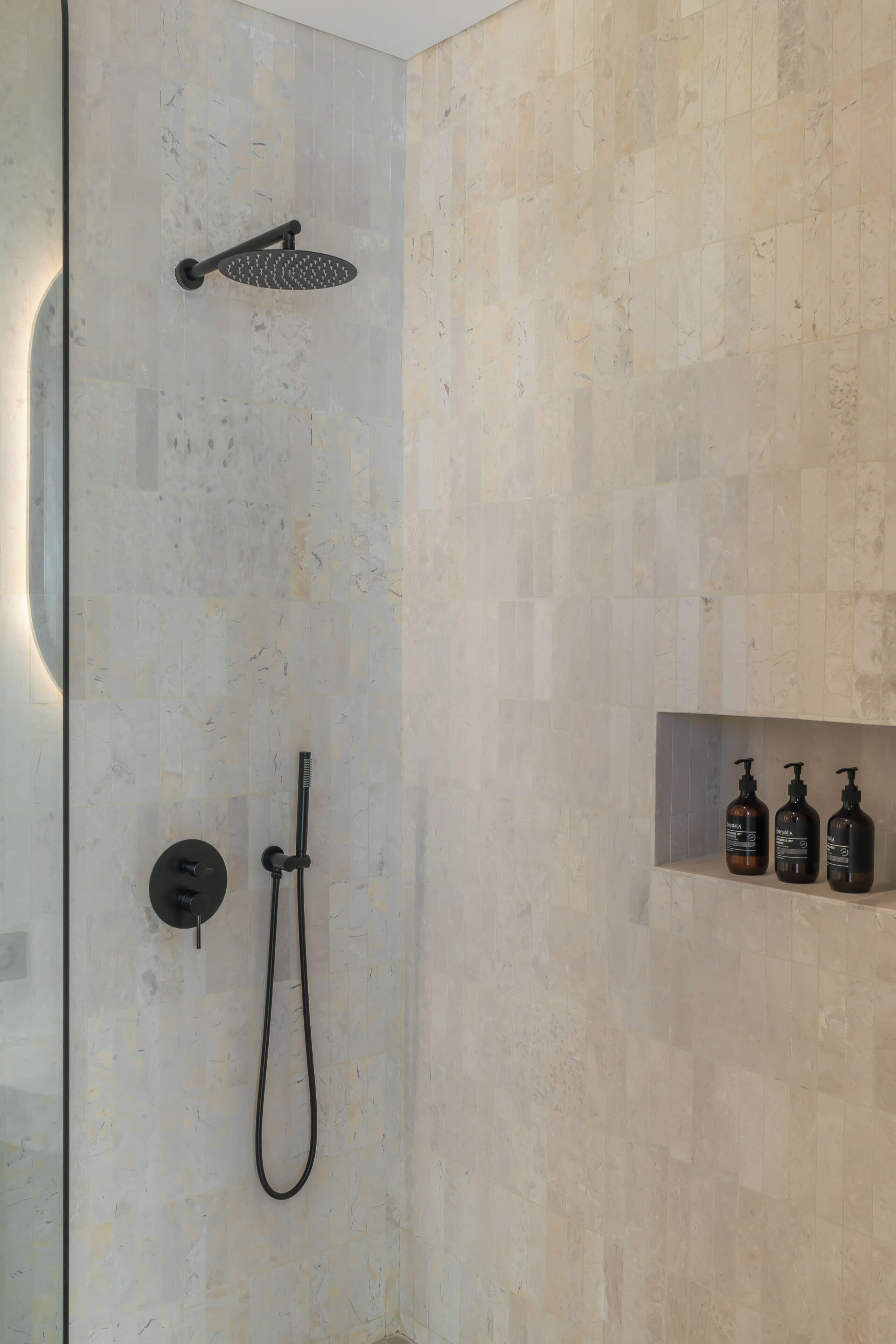 Zona de ducha con cabezal de ducha manual, cabezal de ducha instalado en la pared y manguera para ducha manual. Hay una repisa con tres botellas de champú.