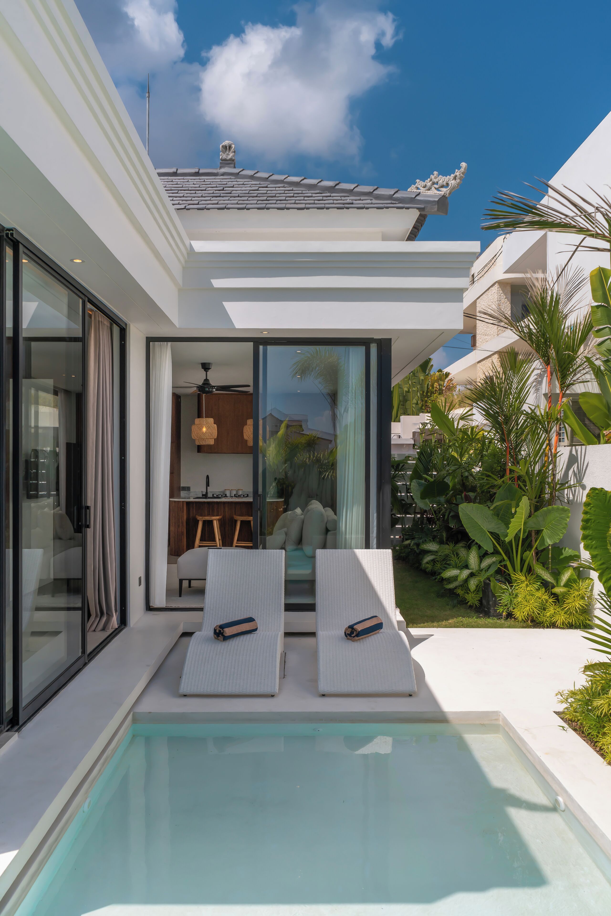 A modern outdoor pool area with two lounge chairs, a glass-enclosed kitchen, and a view of the sky.