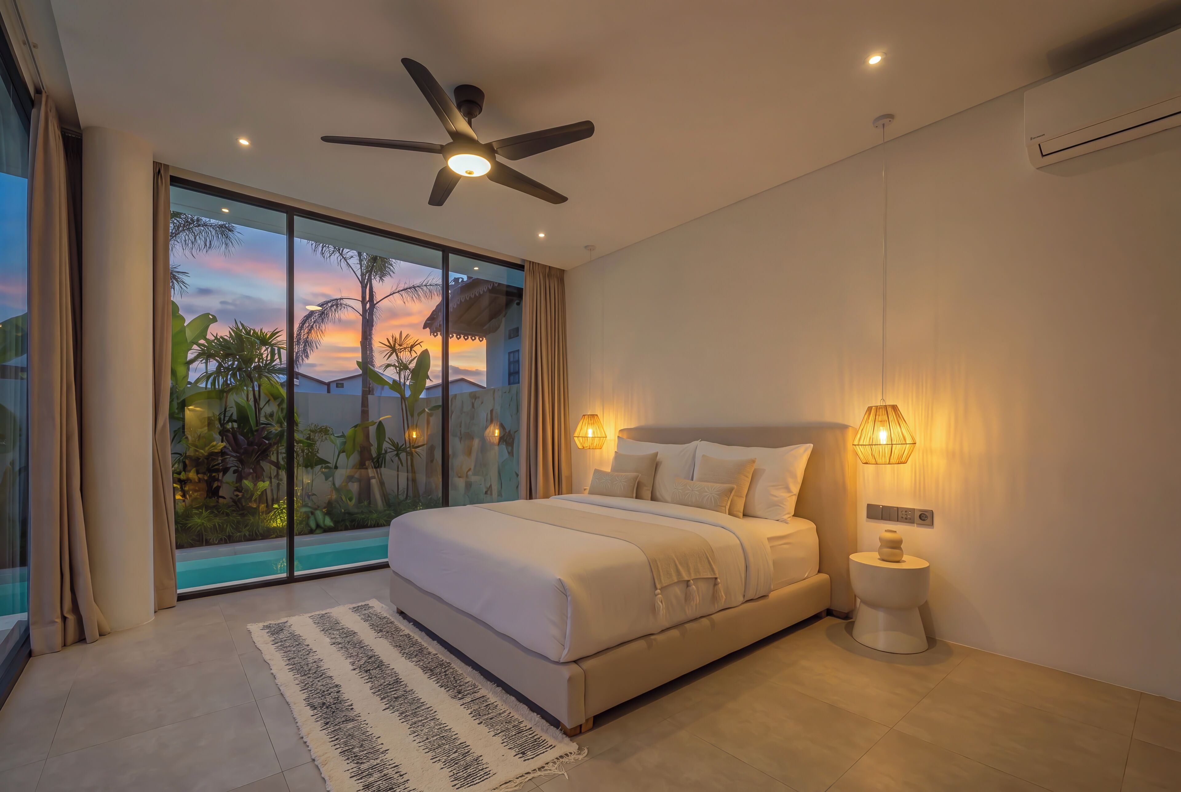 A bedroom with a large bed, a ceiling fan, a nightstand, and a view of palm trees through a glass door.