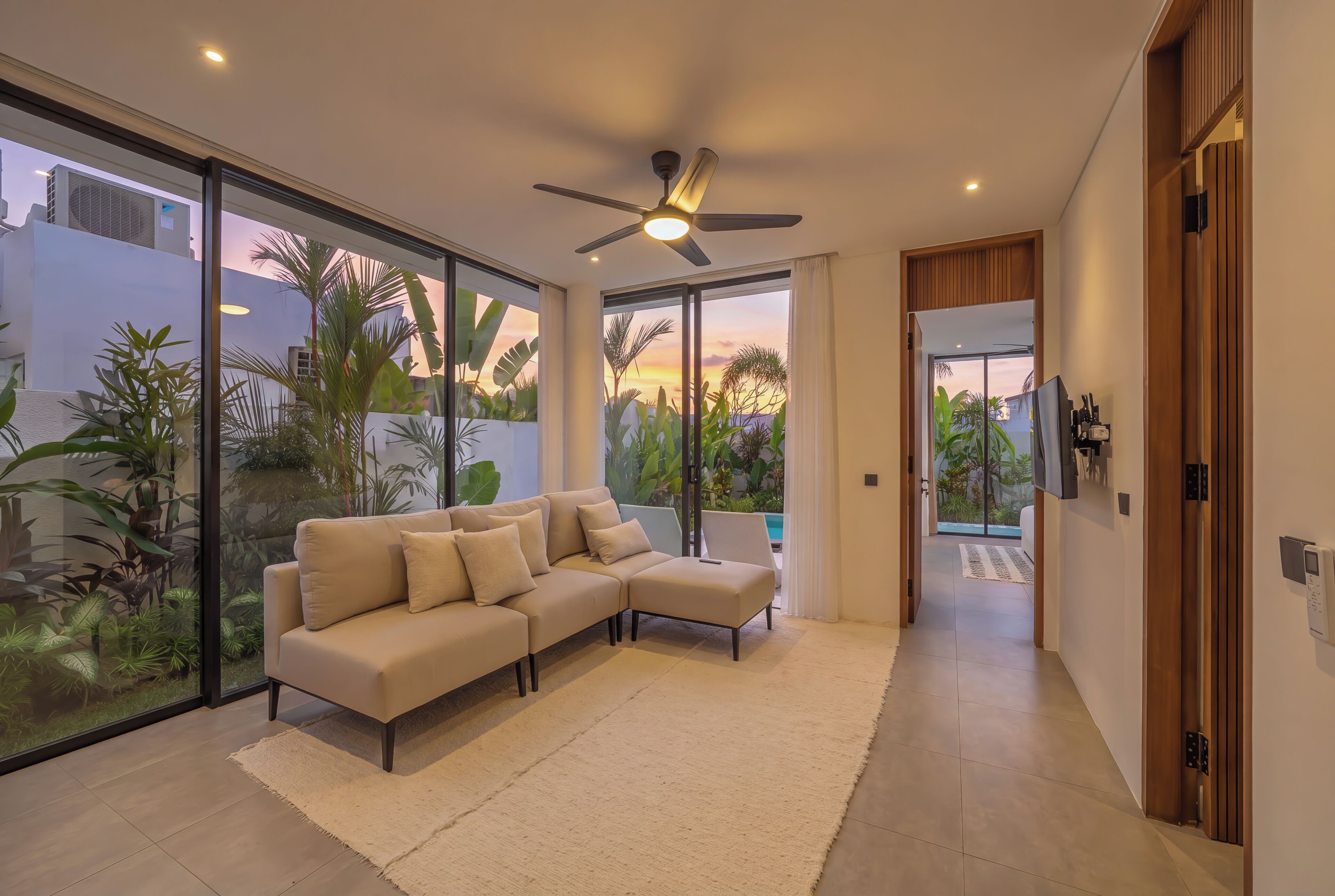 Un salon moderne avec un canapé d’angle, un ventilateur de plafond et de grandes portes-fenêtres coulissantes donnant sur un balcon agrémenté de plantes tropicales.