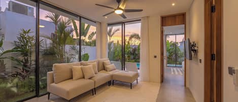 A modern living room with a sectional sofa, a ceiling fan, and large sliding glass doors leading to a balcony with tropical plants.