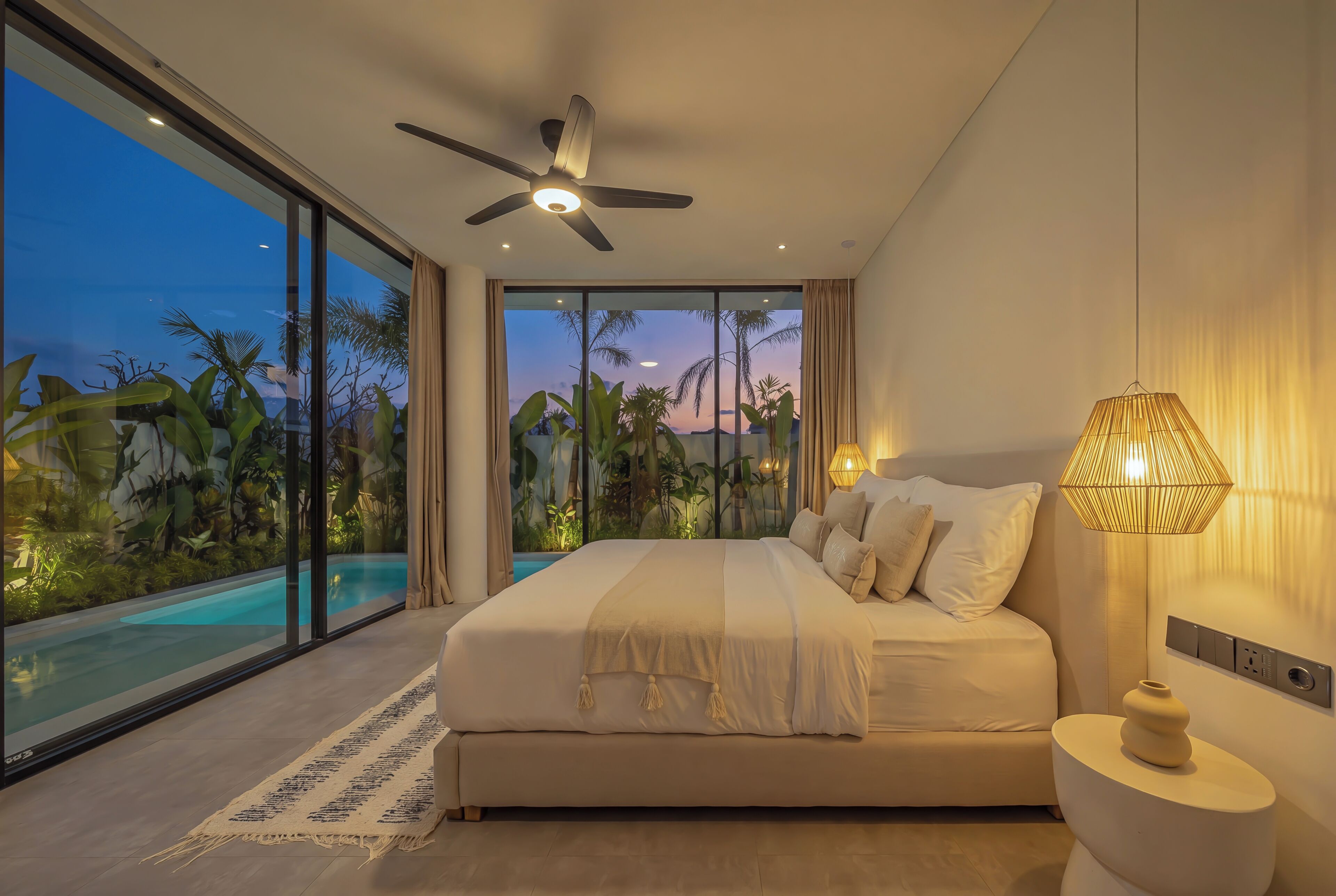 Un dormitorio con una cama grande, ventilador de techo, lámpara de noche y vista a una piscina y plantas tropicales.