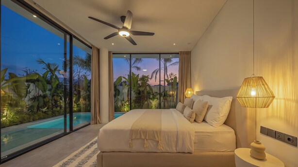 Una camera da letto con un letto grande, un ventilatore a soffitto, una lampada da comodino e vista su una piscina e piante tropicali.