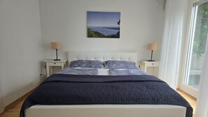 Un dormitorio con una cama grande, dos mesitas de noche, una mesita auxiliar, una ventana con cortinas y un cuadro colgado en la pared.
