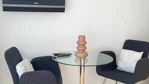 Una habitación con una mesa redonda de vidrio, dos sillas negras, un control remoto y un televisor de pantalla plana montado en la pared.