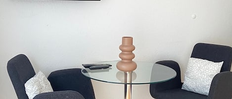 A room with a round glass table, two black chairs, a remote control, and a flat-screen TV mounted on the wall.