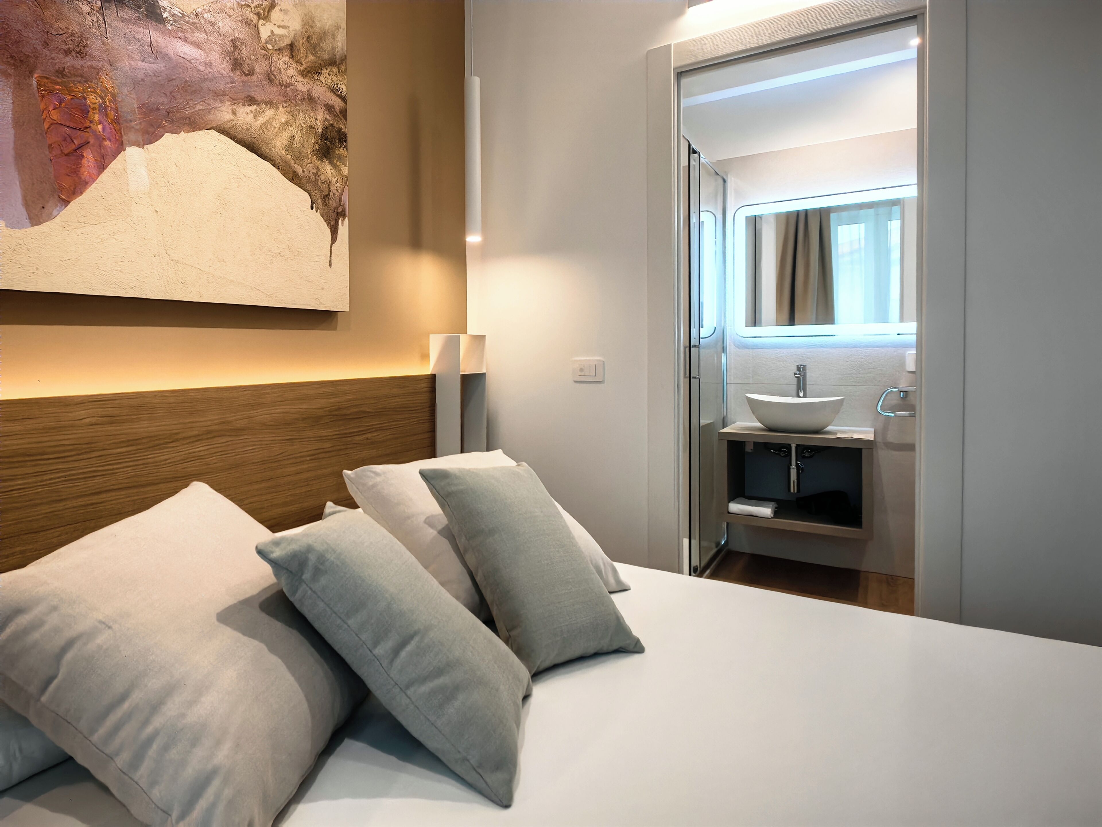 A hotel room with a bed, pillows, a nightstand, a wall-mounted lamp, and a bathroom visible through an open door.