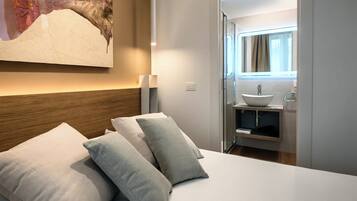 A hotel room with a bed, pillows, a nightstand, a wall-mounted lamp, and a bathroom visible through an open door.