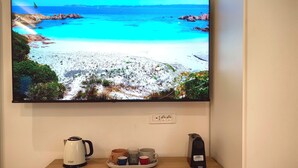 Un televisor montado en la pared muestra una escena de playa. Debajo del televisor, hay una mesa de madera con una tetera, una bandeja con tazas y una cafetera.