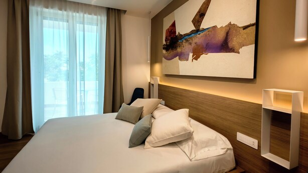 Habitación de hotel con una cama grande, cabecera con iluminación integrada, un cuadro abstracto colgado en la pared y vistas al exterior a través de cortinas traslúcidas.