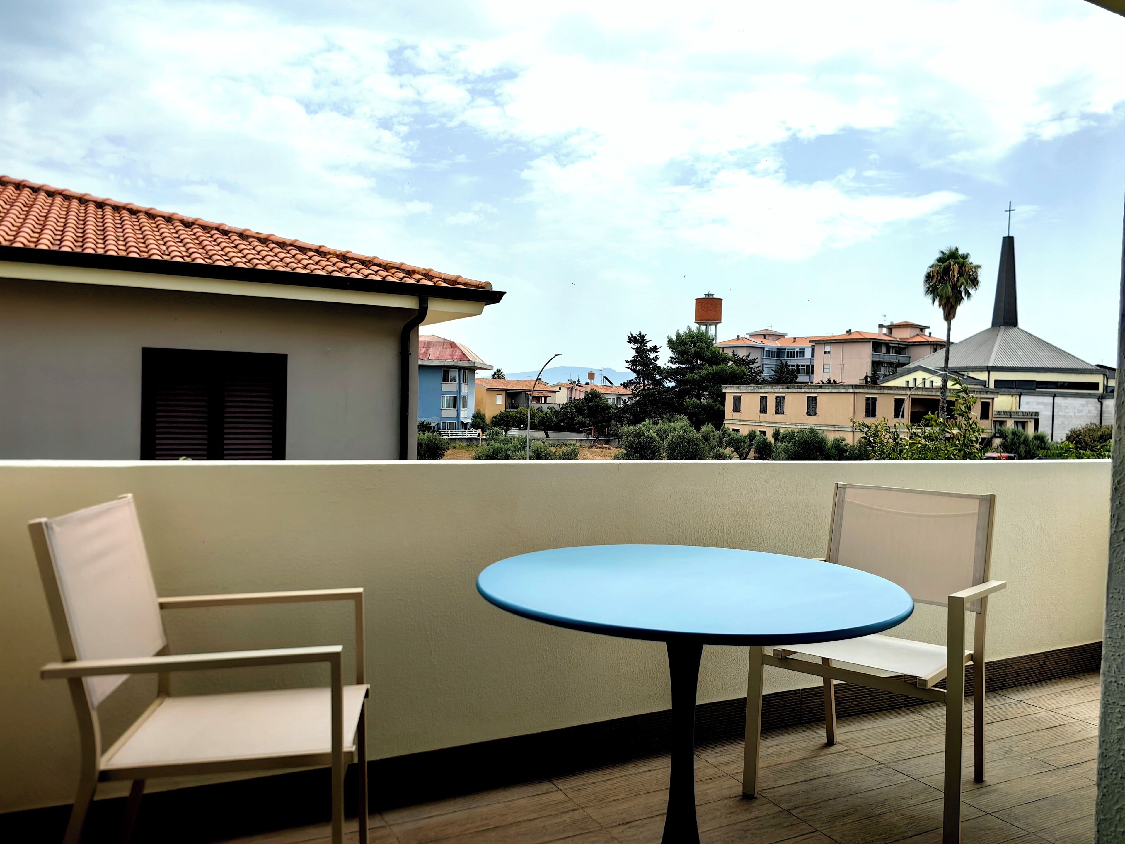 Ein Balkon mit zwei Stühlen und einem Tisch, der auf einen Wohnbereich mit Gebäuden und einer Kirche blickt.