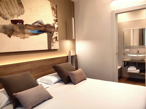 A hotel room with a large bed, a nightstand, a wall-mounted lamp, and a bathroom with a sink and mirror.