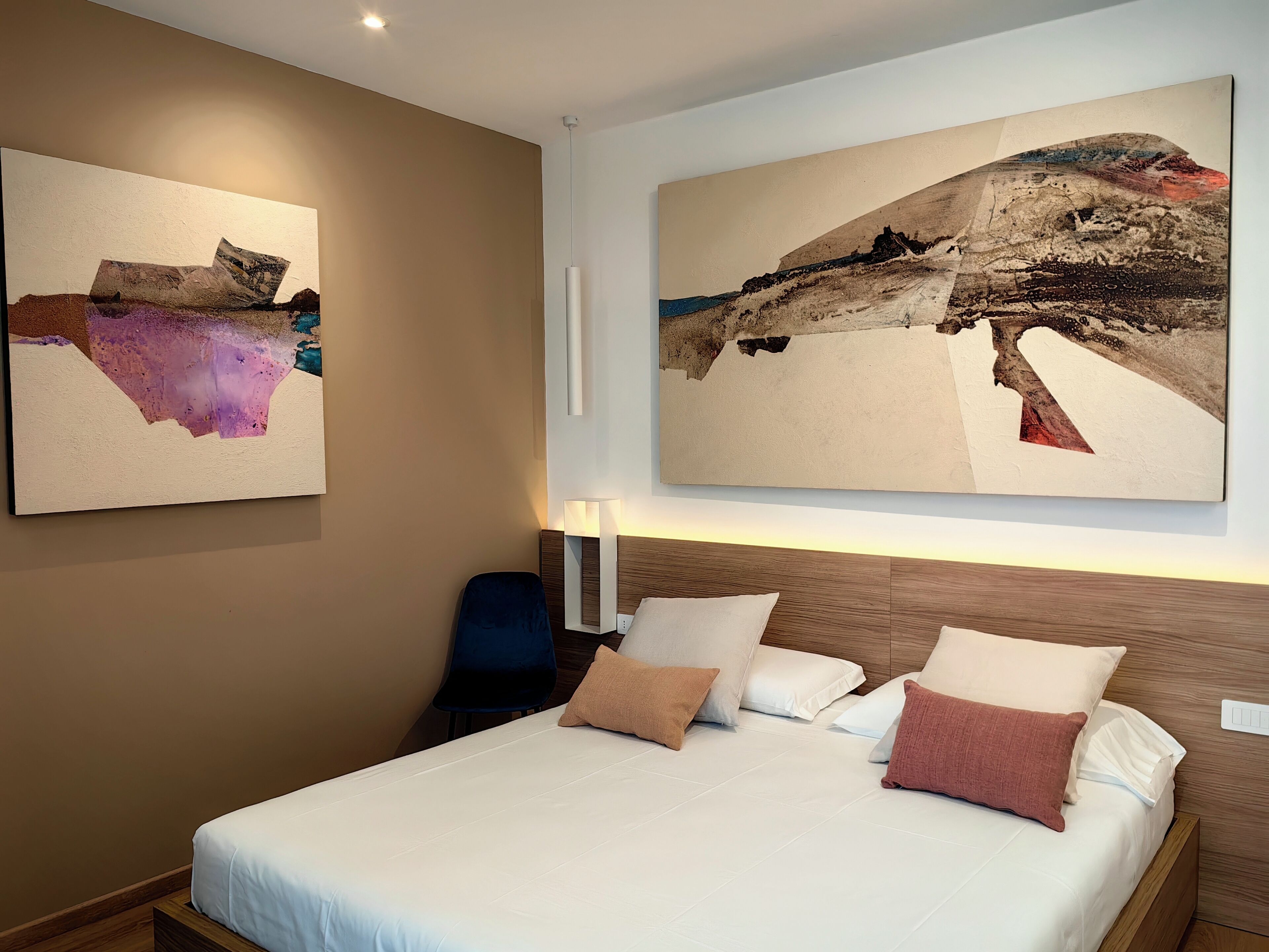 A hotel room with a bed, pillows, a chair, and two large abstract paintings on the wall.