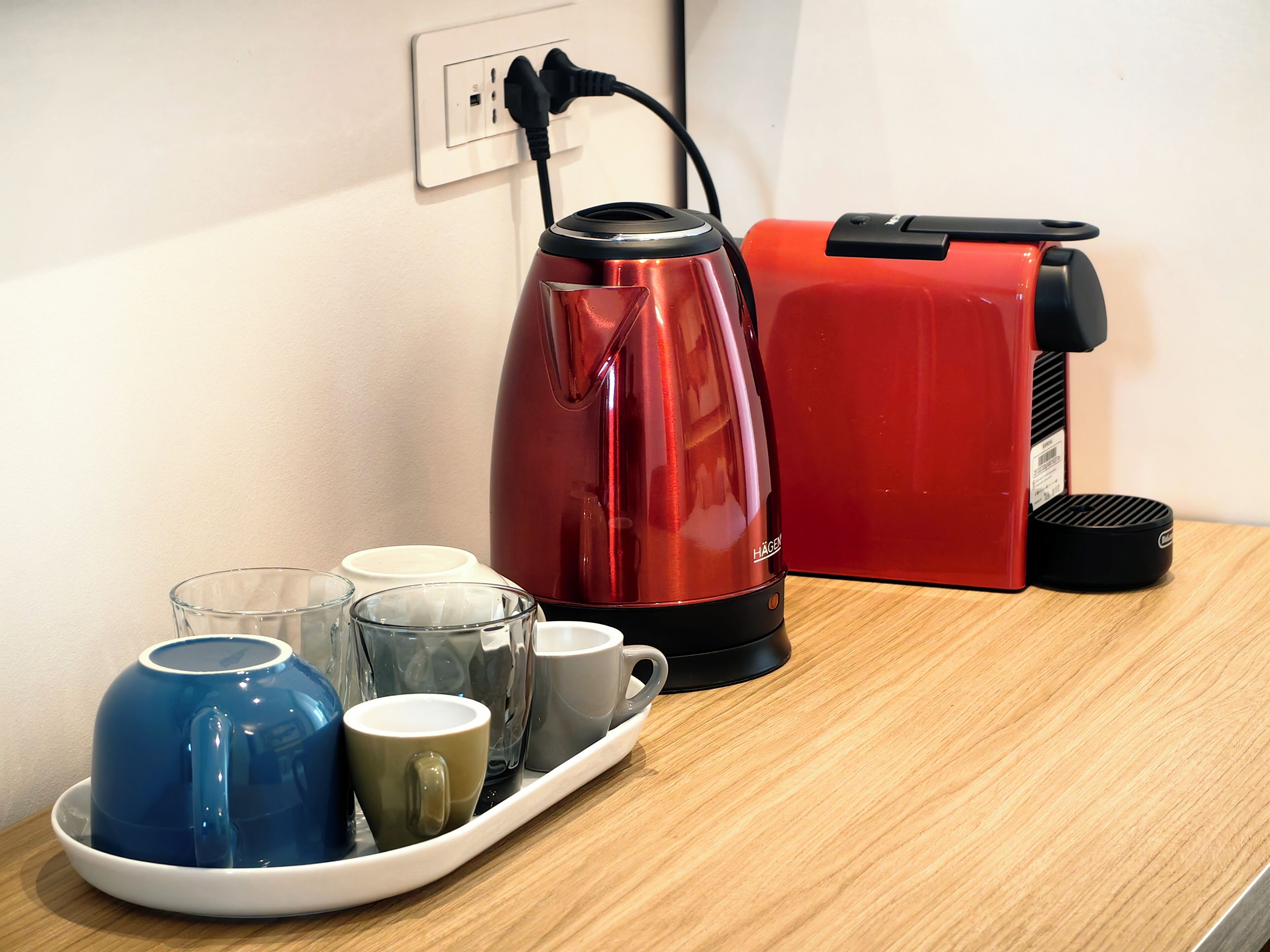 A kitchen area with a red electric kettle, a red coffee maker, and a tray with various cups and a blue mug.