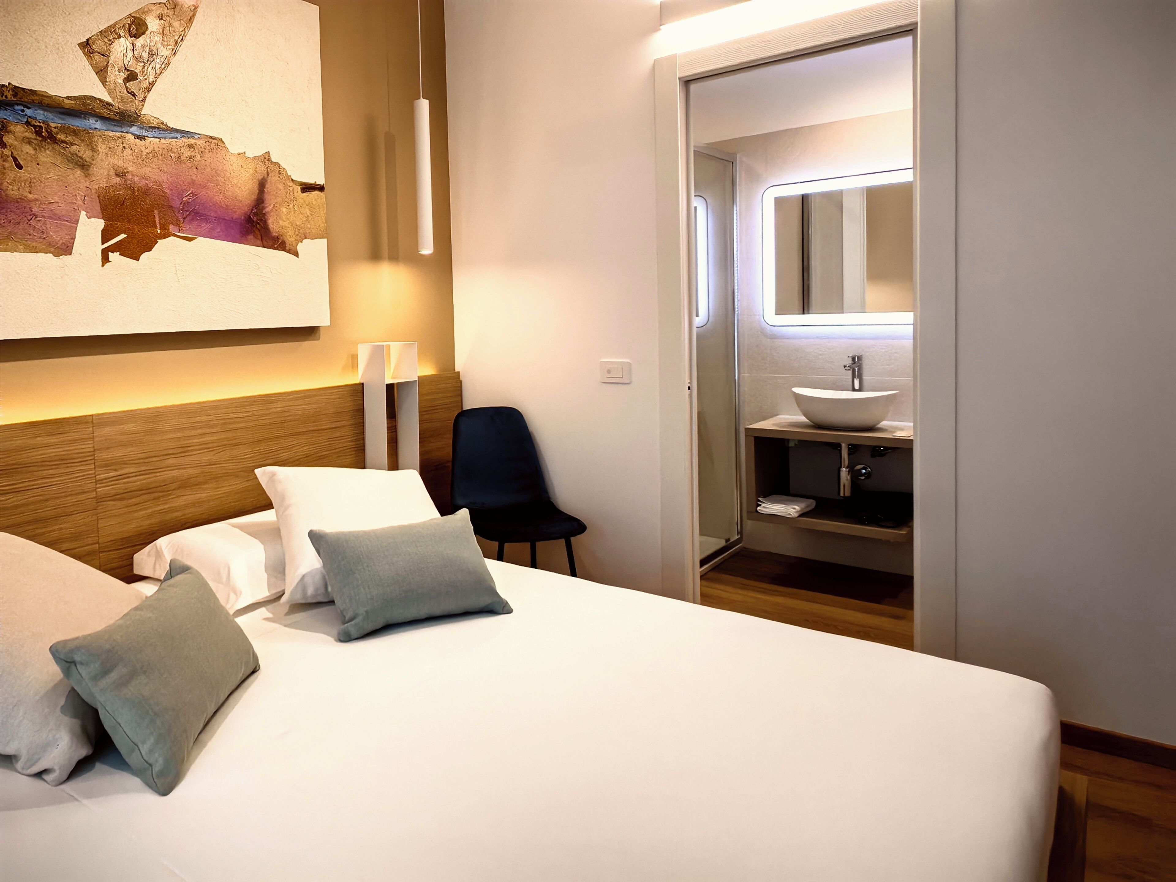 A hotel room with a bed, pillows, a chair, a bedside table, a wall-mounted lamp, and a bathroom with a sink and mirror.