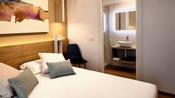 A hotel room with a bed, pillows, a chair, a bedside table, a wall-mounted lamp, and a bathroom with a sink and mirror.