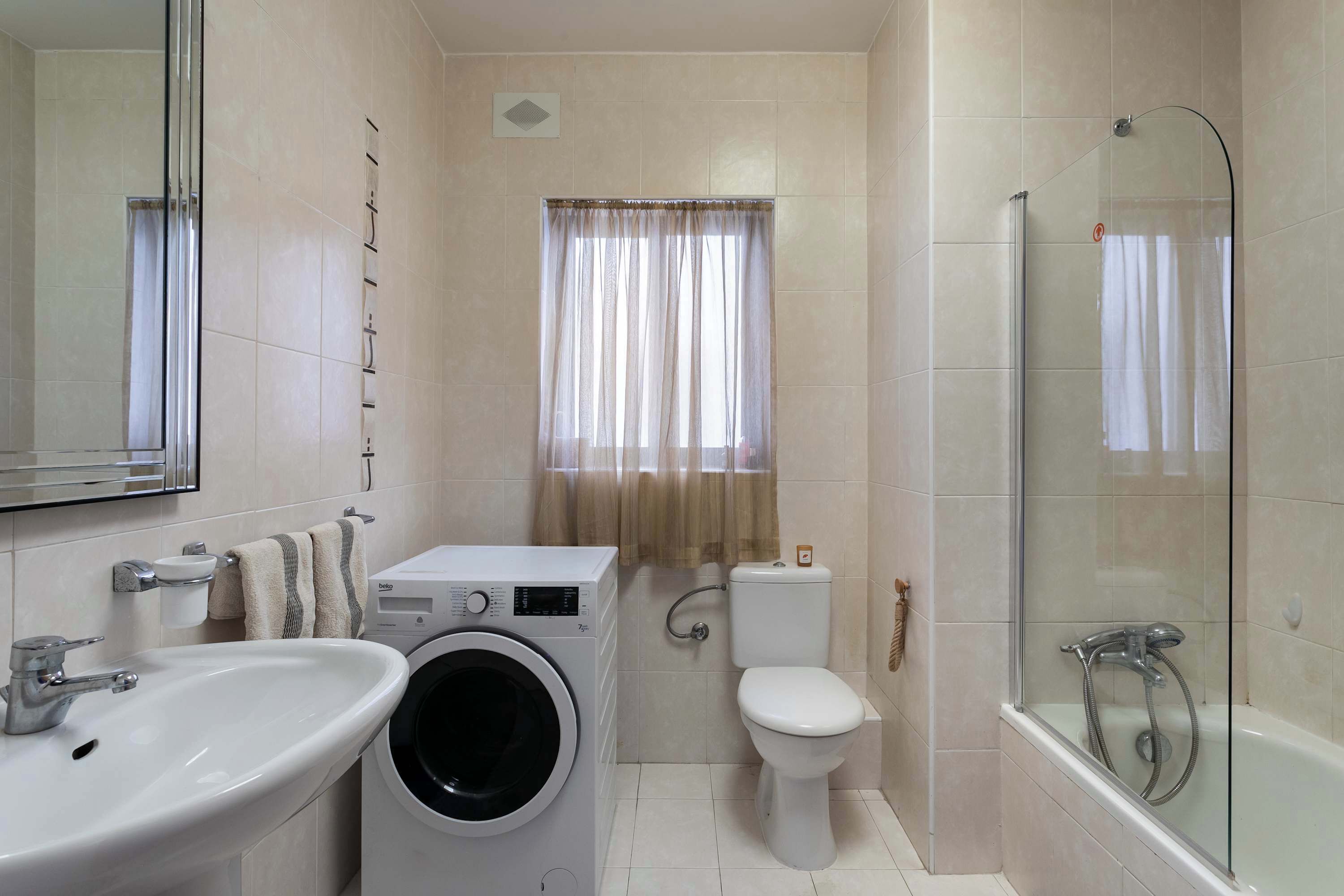 A bathroom with a sink, washing machine, toilet, and glass-enclosed shower.
