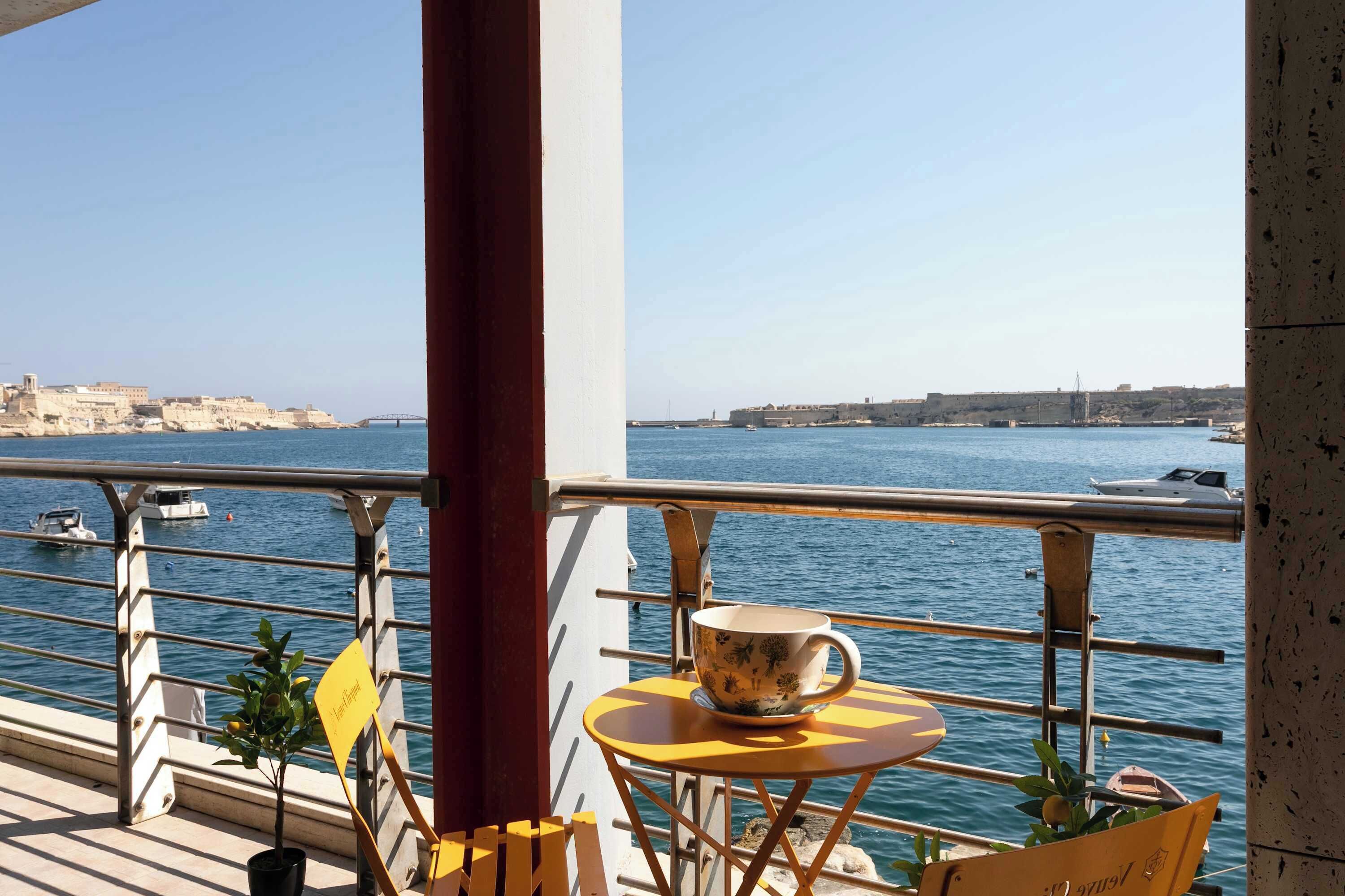 A balcony with a table and chairs overlooking a body of water, with buildings in the background.