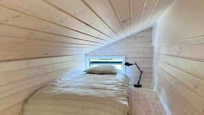 Ein kleiner Raum mit schrägem Dach, einem Bett, einem Fenster und einer Stehlampe.