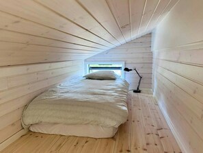 Ein kleiner Raum mit schrägem Dach, einem Bett, einem Fenster und einer Stehlampe.