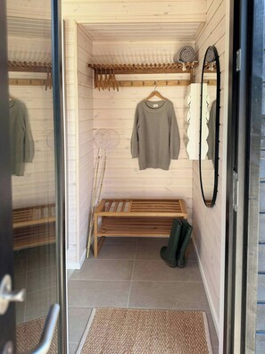 A small, enclosed room with a bench, a hanging rack with clothes, a mirror, and a pair of boots.