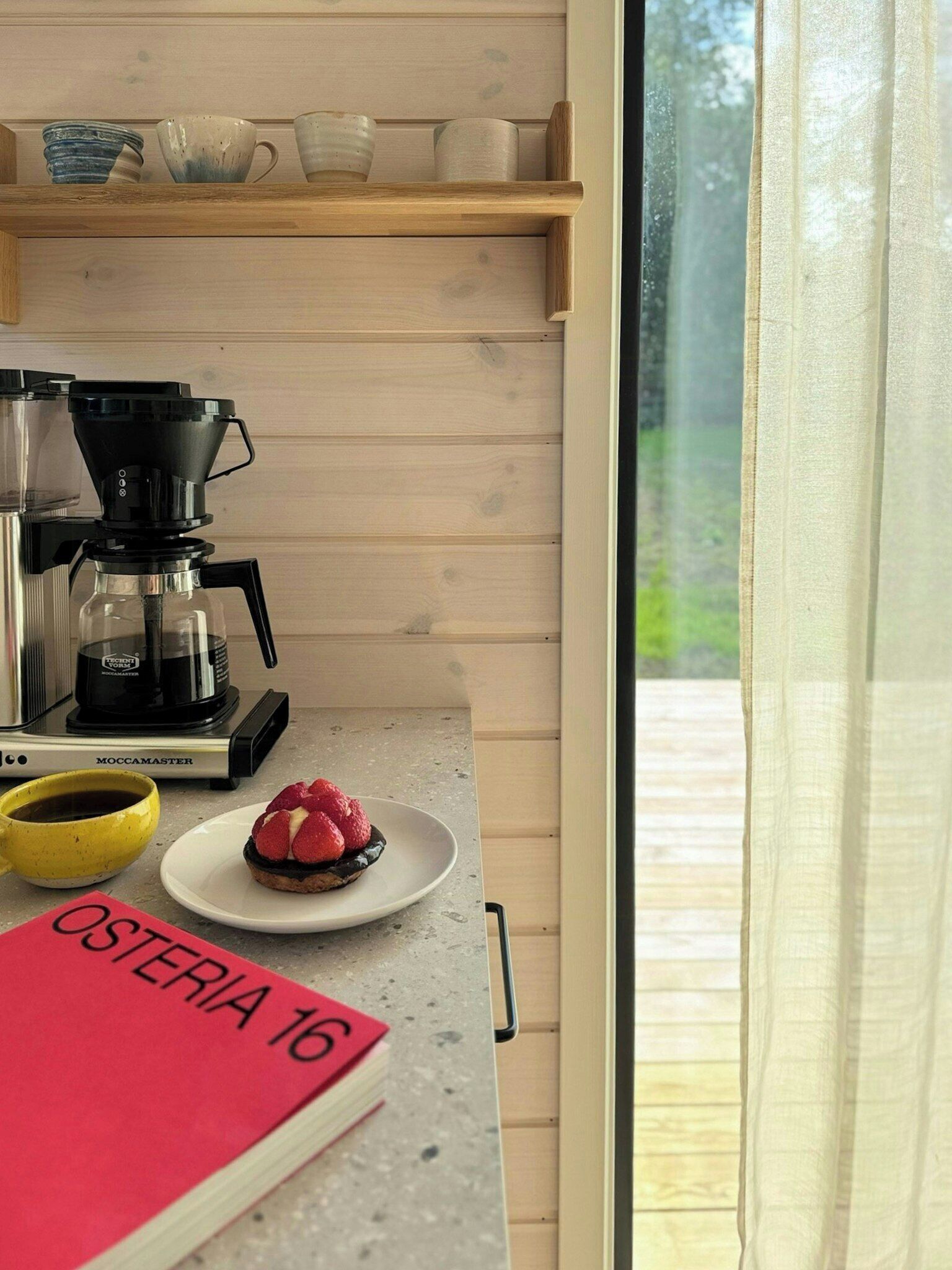 A kitchenette with a coffee maker, a plate with a dessert, a yellow mug, and a red sign with 'OSTERIA 16' on a countertop.