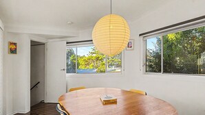 Una sala menjador amb taula de fusta, cadires, llum penjant i finestres amb vista a arbres.