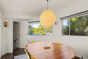 A dining room with a wooden table, chairs, a hanging light fixture, and windows with a view of trees. - Coastal Nostalgia – Classic charm, Coastal comfort (Macmasters Beach)