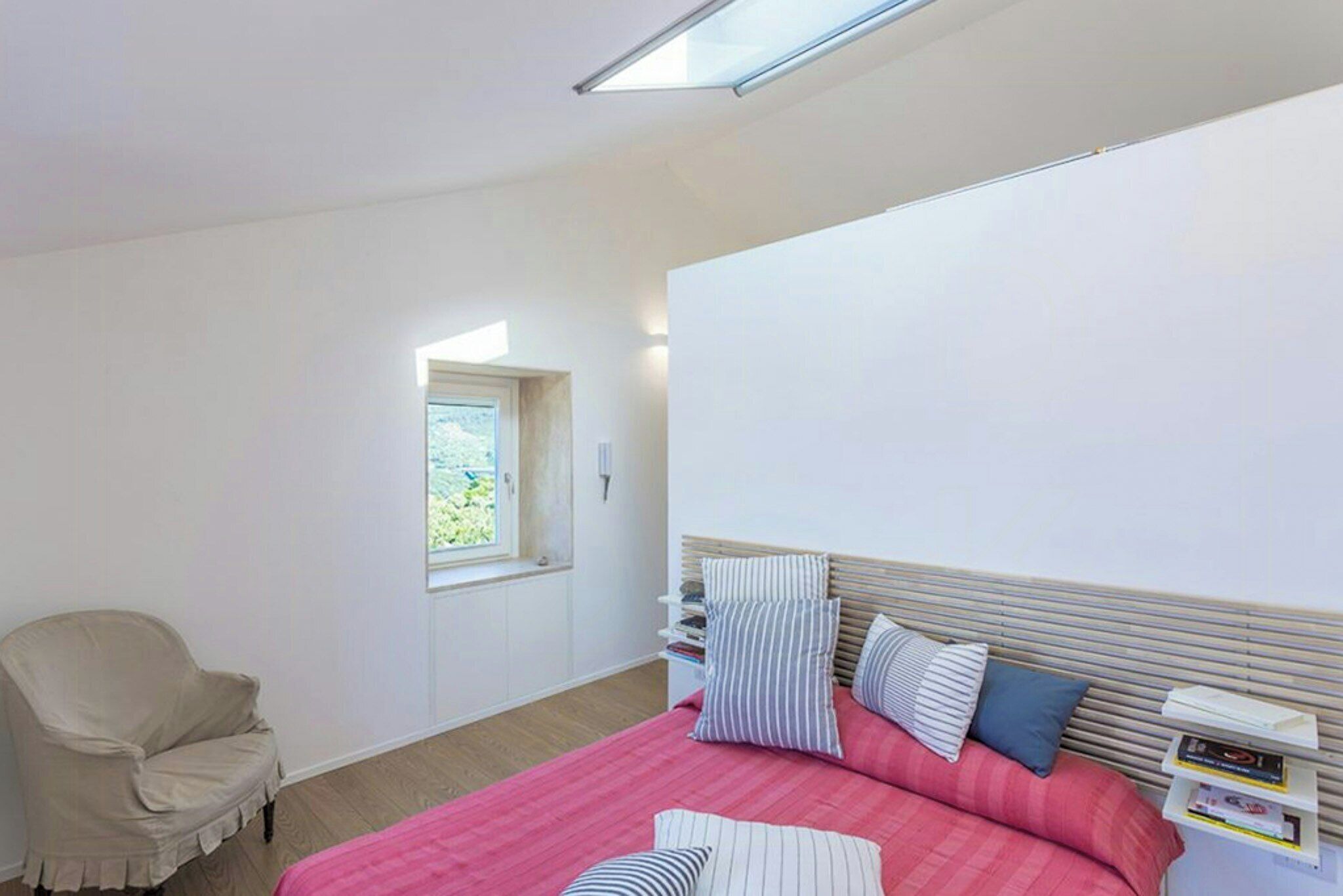 A bedroom with a bed, a chair, a window, and a bedside table with books.
