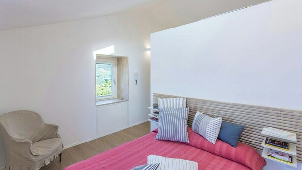 Ein Schlafzimmer mit einem Bett, einem Sessel, einem Fenster und einem Nachttisch mit Büchern.