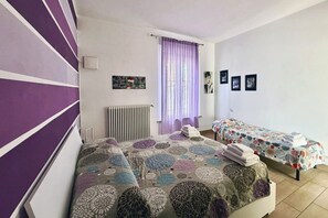 A room with two beds, a radiator, a window with curtains, and a wall with framed pictures. - Spacious 89 m² apartment, ideal for 6 people, located on the ground floor of a period building with a private entrance (accessible via stairs).Situated in the heart of the historic center, the property offers a splendid view of Rocca Brancaleone Park an (Ravenna)