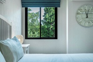 A bedroom with a bed, a bedside table, a window with a view of trees, and a wall clock. - The Title Residencies Naiyang Beach Apartment E413 (Thalang/Sakhu)