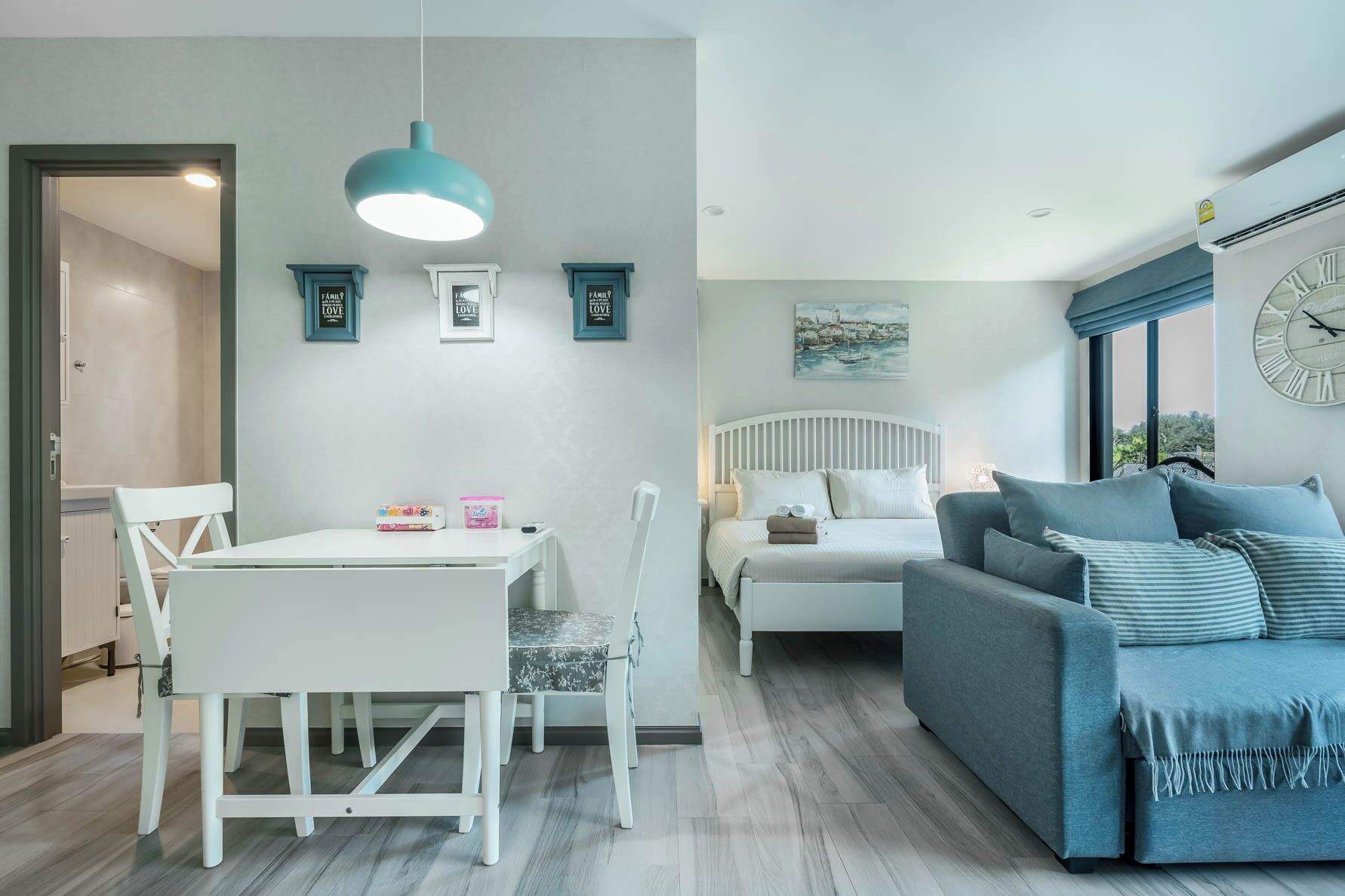 Una sala moderna con una cama blanca, un sofá gris, una mesa de comedor con sillas y una lámpara colgante.