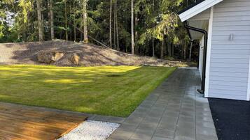 Un patio ajardinado con terraza de madera, un sendero pavimentado y un edificio blanco.