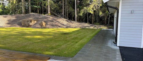 A landscaped yard with a wooden deck, a paved walkway, and a white building.
