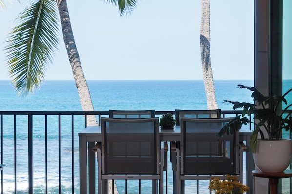 A balcony with two chairs facing the ocean, palm trees, and a potted plant.