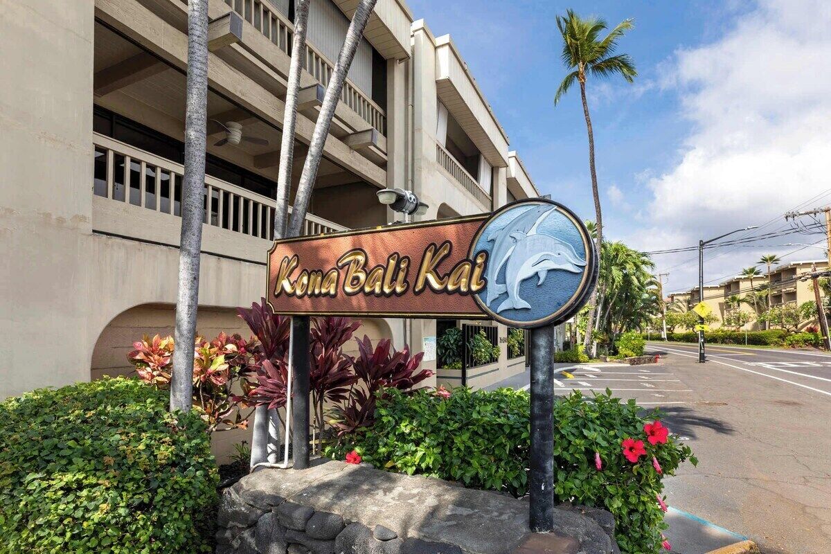 A sign for Kona Bali Kai with a dolphin logo, surrounded by tropical plants and a building in the background.