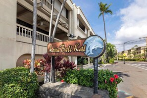 A sign for Kona Bali Kai with a dolphin logo, surrounded by tropical plants and a building in the background. - Beautiful Completely Renovated 2 Bed/2 Bath Oceanfront Gem - Kona Bali Kai #313 (Kailua-Kona)