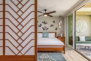 A bedroom with a wooden bed, a ceiling fan, decorative wall art, a bedside table with a lamp, and a view of a green sofa through a glass door.
