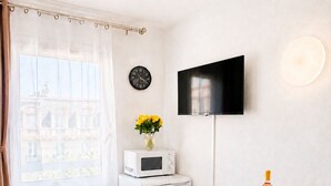 A compact living space with a dining area, a microwave, a flat-screen TV, a clock, and a window with a curtain. - Le Thiers Azur AP4520 by Riviera Holiday Homes (Nice)