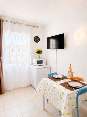 A compact living space with a dining area, a microwave, a flat-screen TV, a clock, and a window with a curtain. - Le Thiers Azur AP4520 by Riviera Holiday Homes (Nice)