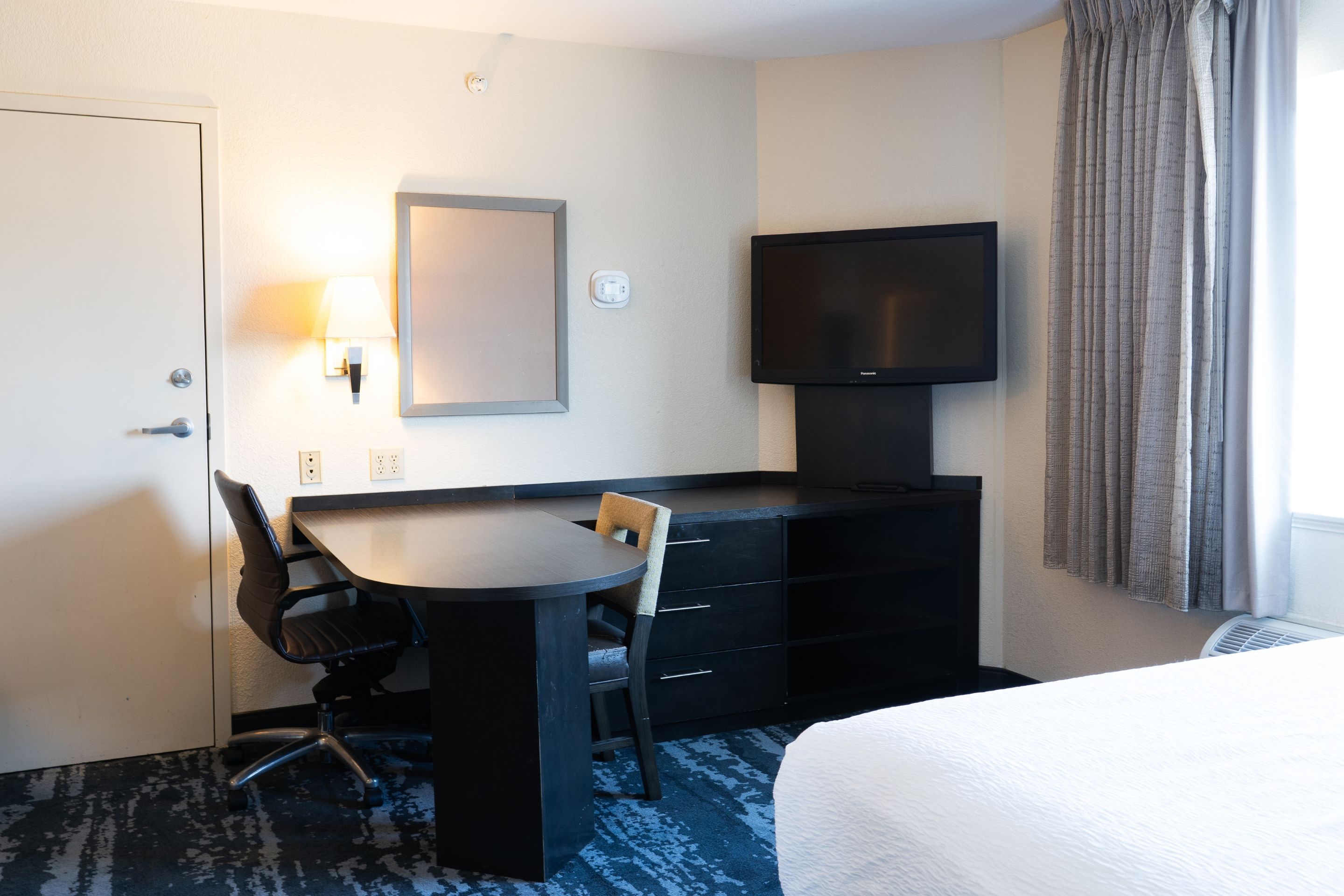 Una habitación de hotel con una cama, un escritorio con silla, un televisor y un espejo.