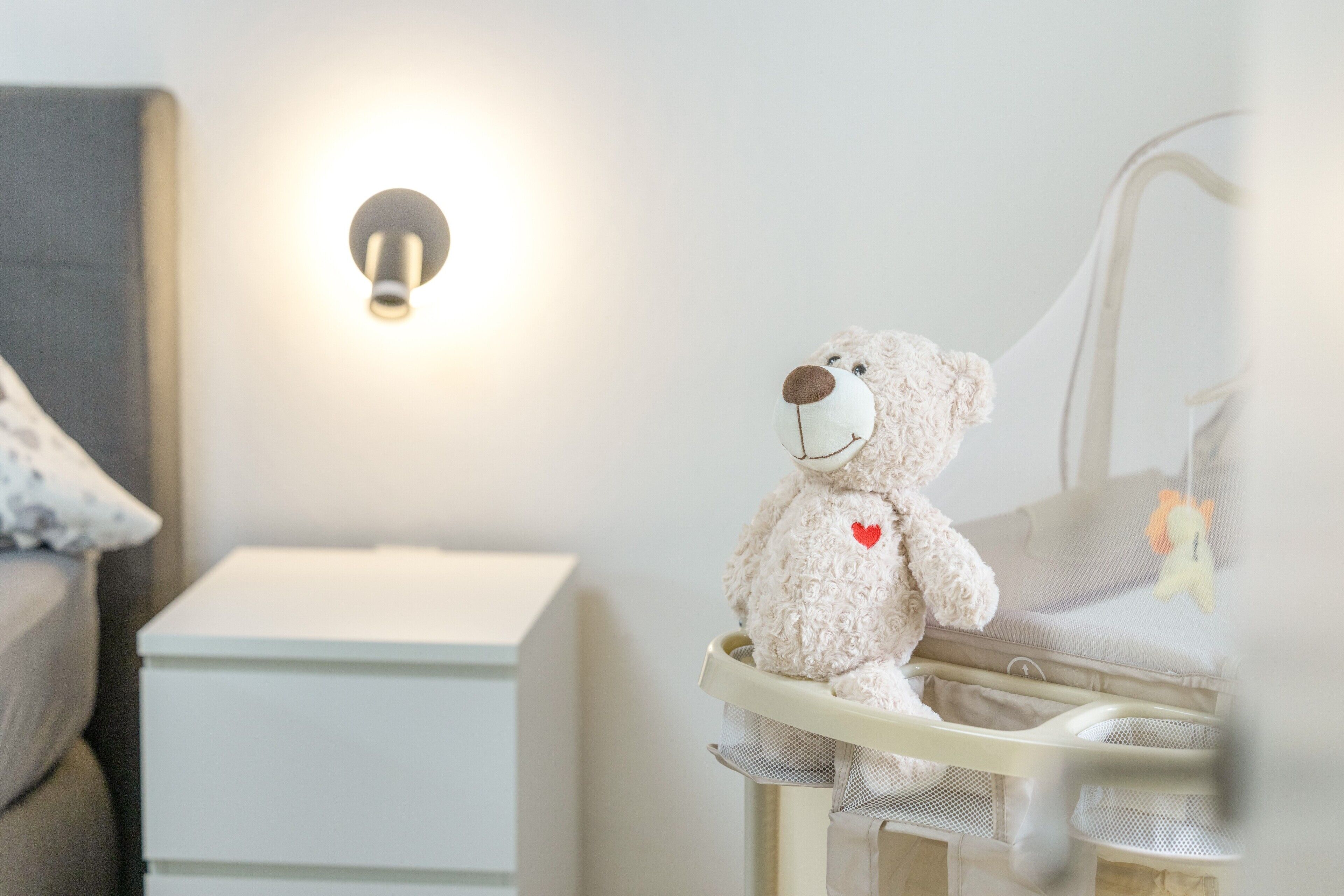 Ein Babybett mit Teddybär, ein Nachttisch und eine Wandleuchte.