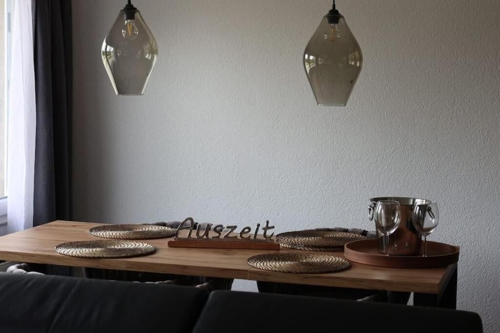 A dining table with woven placemats, a wooden board with the word 'Auszeit,' and two wine glasses on a copper tray.
