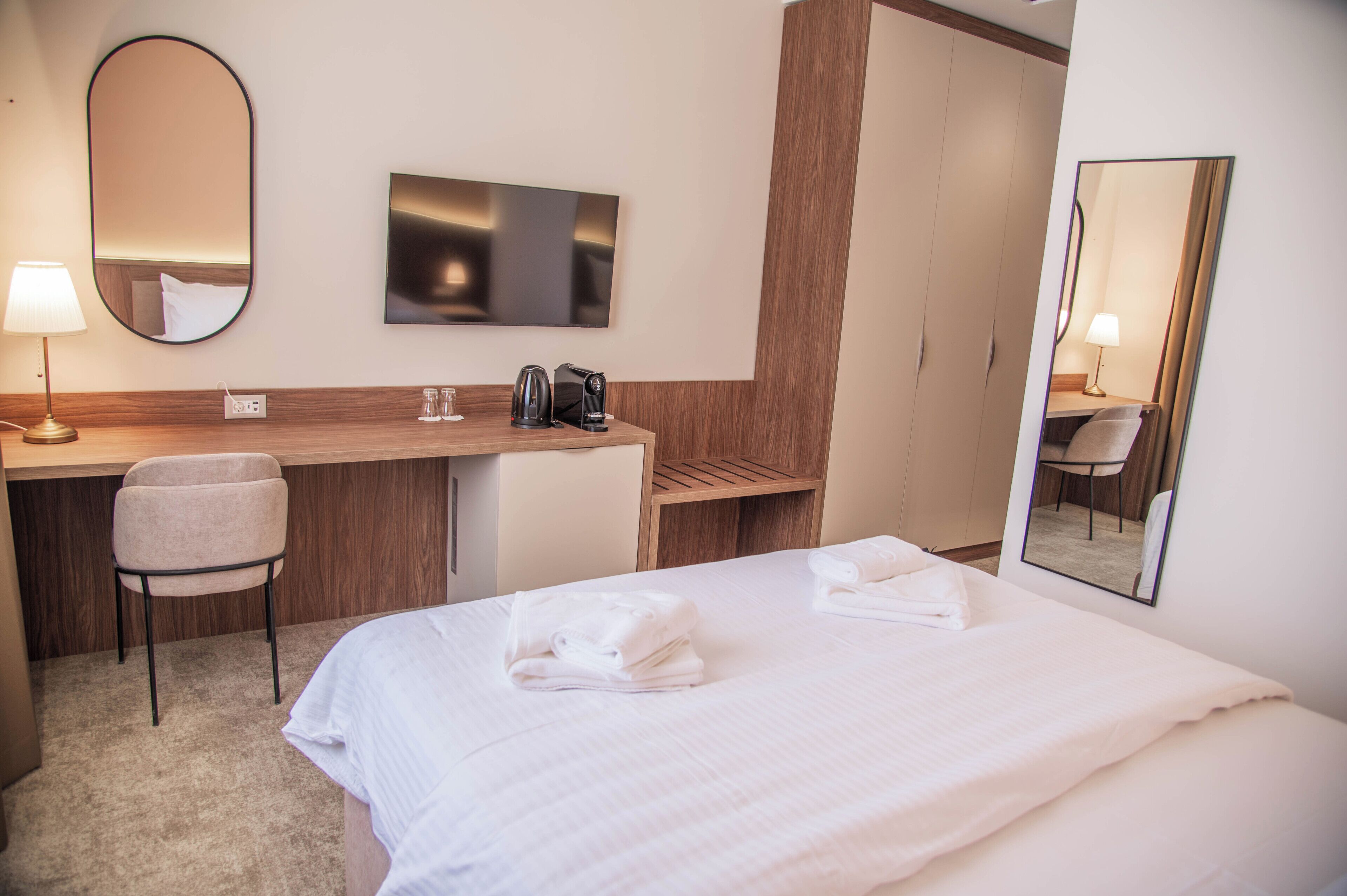 A hotel room with a bed, a desk, a chair, a coffee maker, a mirror, and a television.