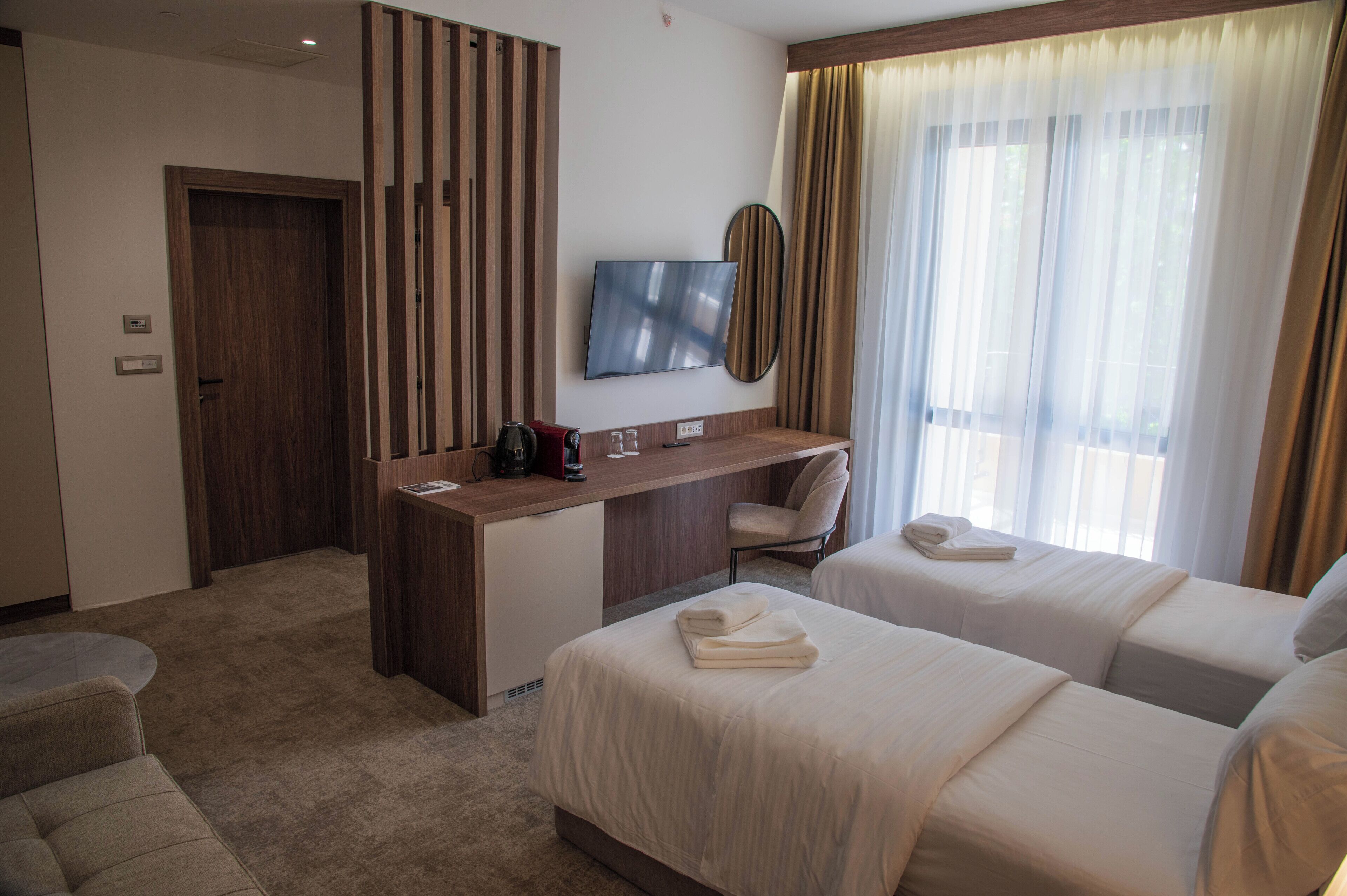 A hotel room with a desk, two beds, a chair, a TV, and a window with curtains.